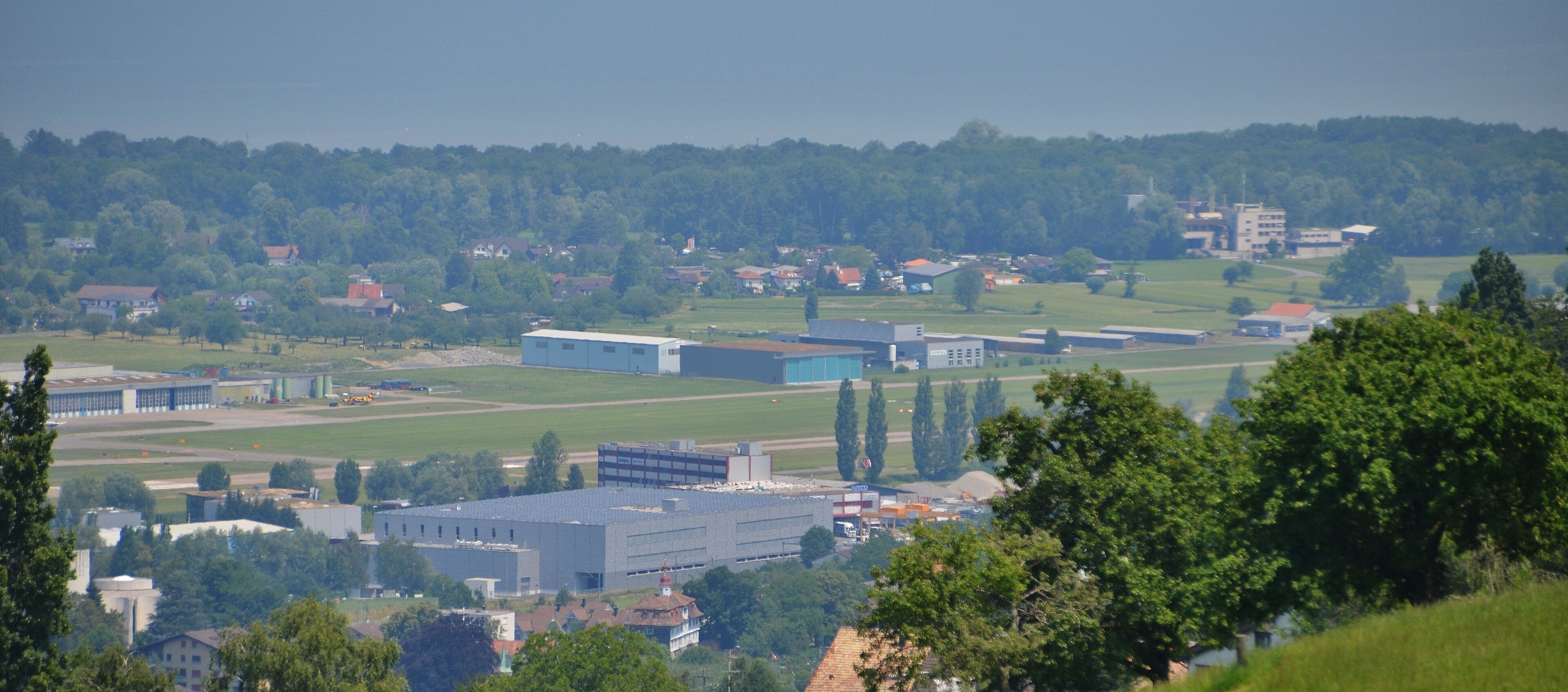 A landscape with industrial buildings, a large blue structure, and a forested hill in the background.