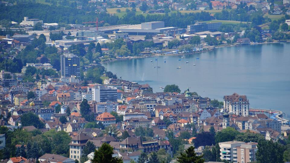 A coastal city with a mix of historic and modern buildings, a large body of water with boats, and a hilly landscape in the background.