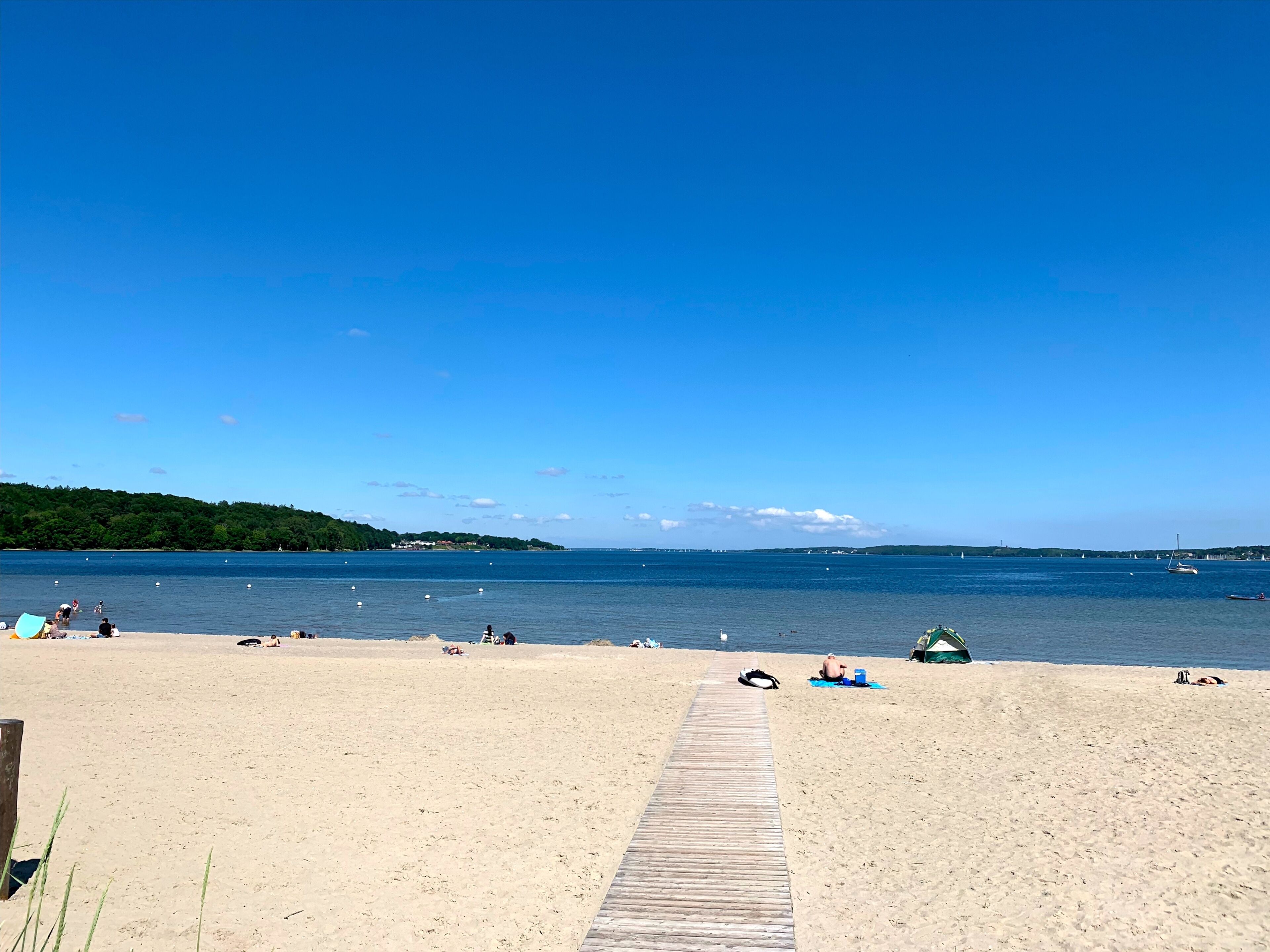A sandy beach with a wooden walkway leading to the water, boats, and a forested island in the distance.
