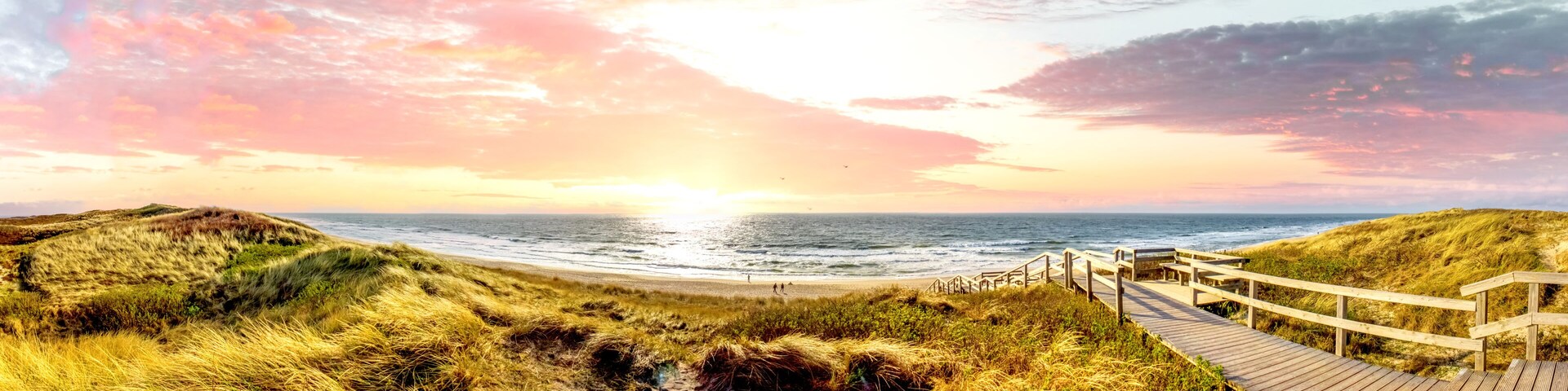 A coastal landscape with dunes, a wooden boardwalk, and the ocean at sunset.