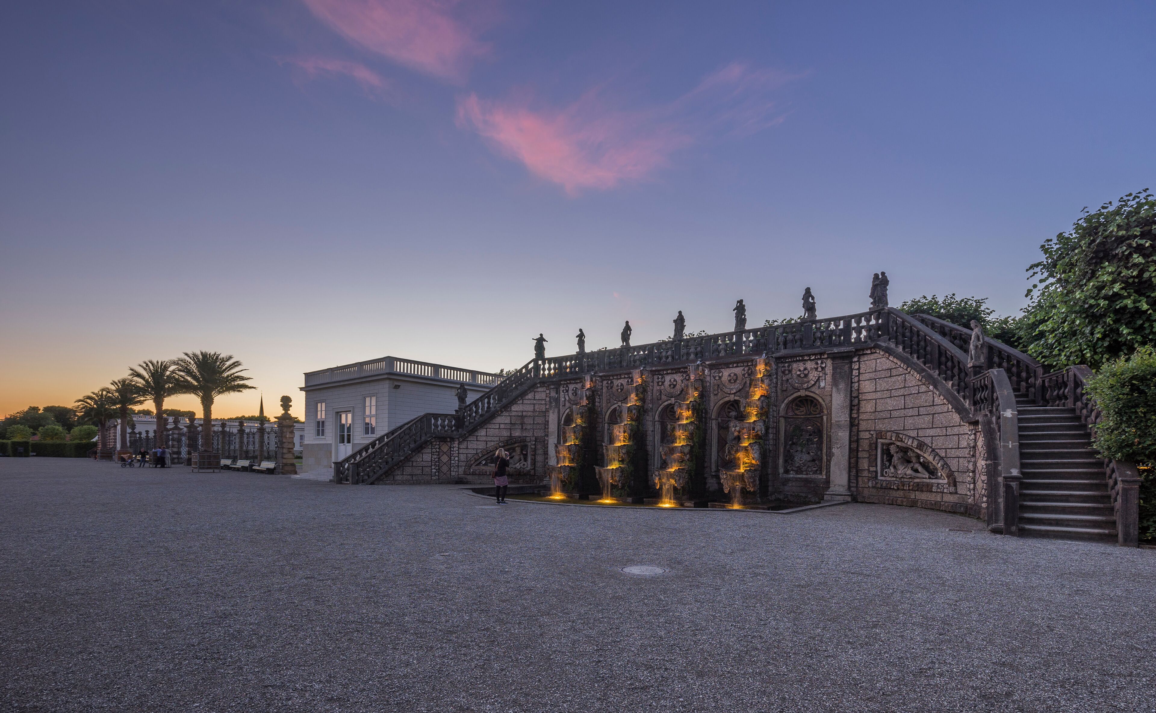 A grand staircase leading to a building with statues, surrounded by a gravel courtyard and palm trees.