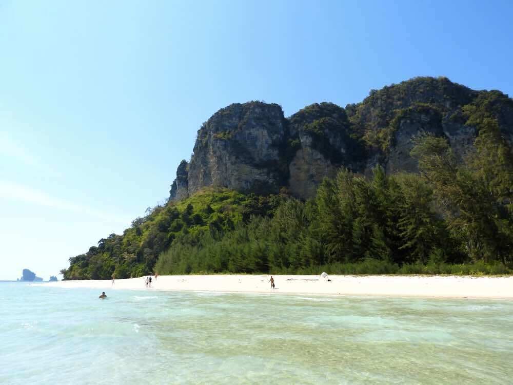 A beach with clear turquoise water, white sand, and a large cliff with dense green vegetation.