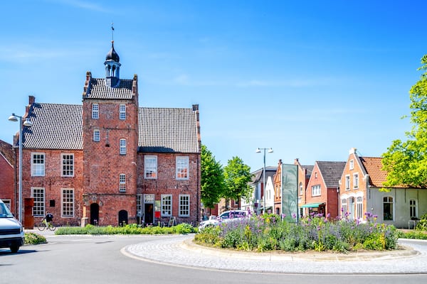 Ein historisches Gebäude aus rotem Backstein mit einem Turm, umgeben von traditioneller europäischer Architektur und einem Pflasterrondell.