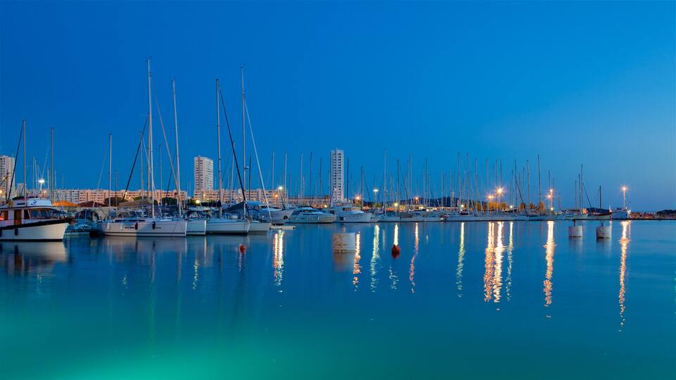 A marina at dusk with numerous sailboats docked, reflecting city lights on the water.