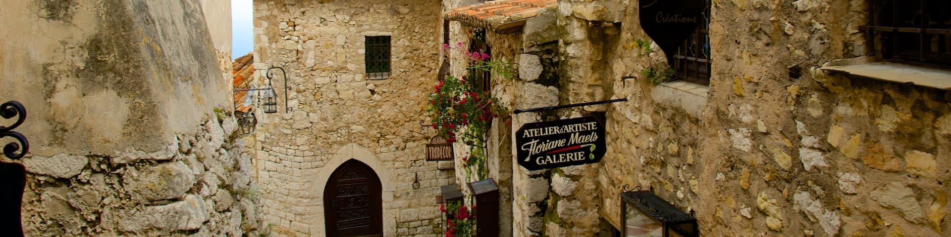 A narrow street with stone buildings, a sign for an artist's gallery, and a bell tower.