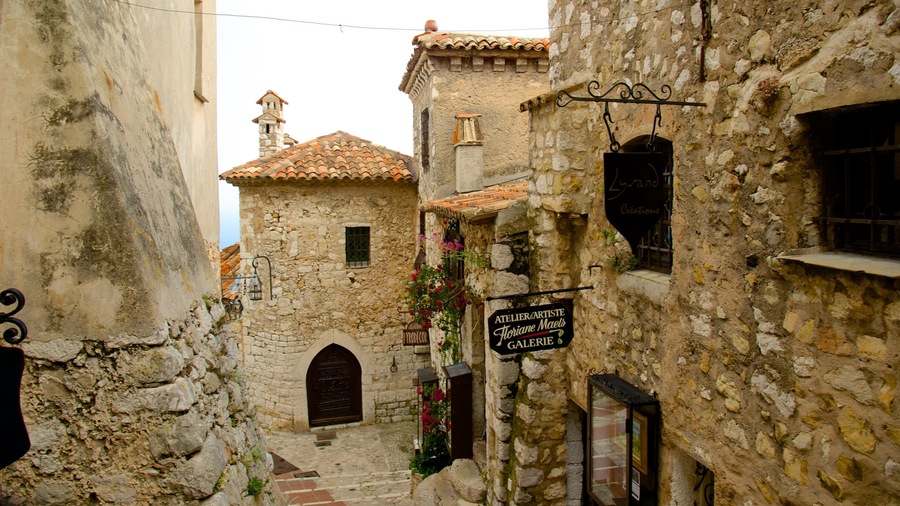 A narrow street with stone buildings, a sign for an artist's gallery, and a bell tower.