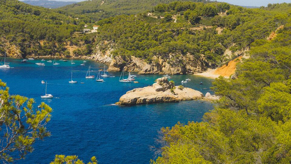 A coastal bay with clear blue water, several sailboats, rocky outcrops, and lush green hills.
