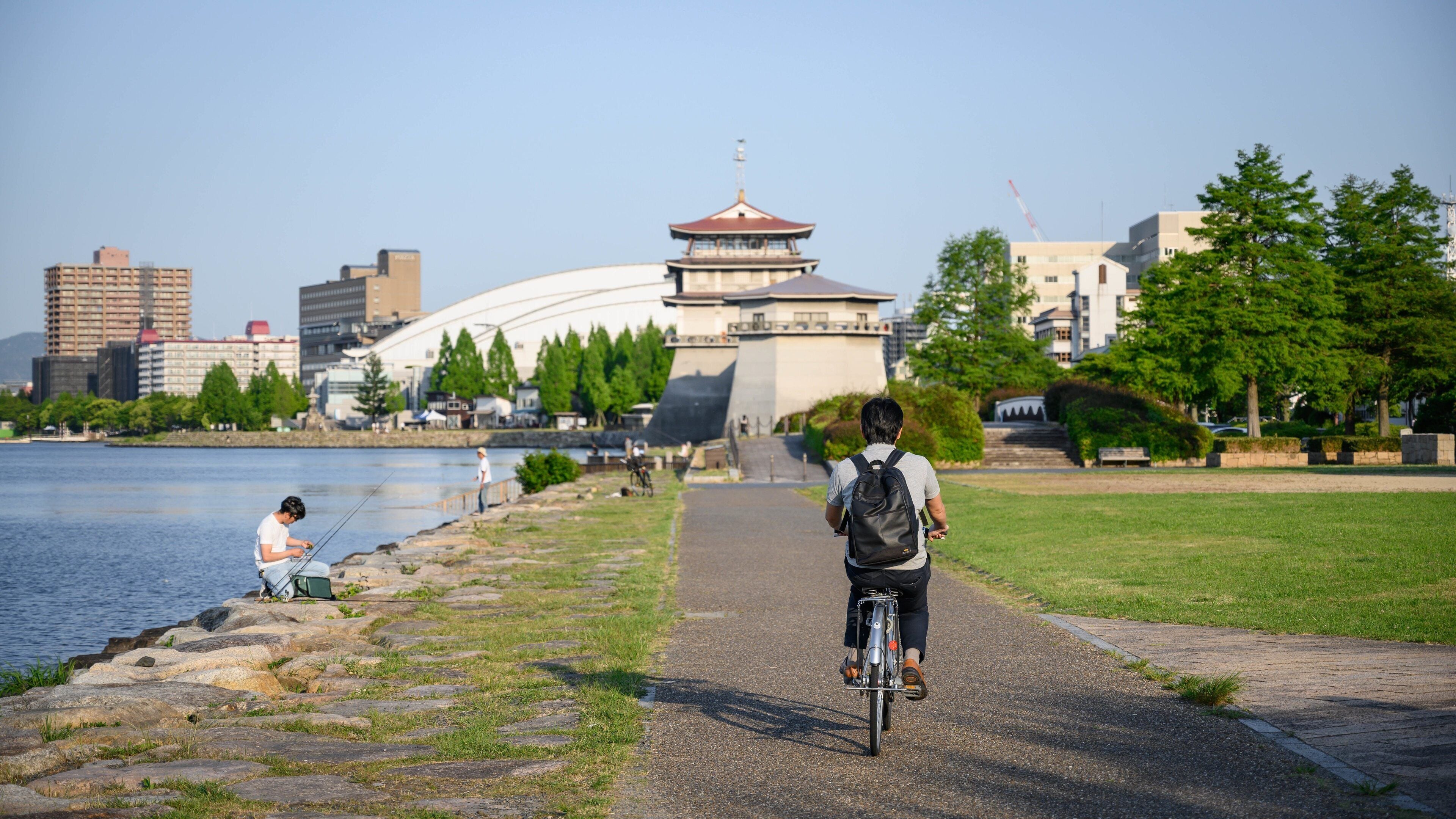 Una persona montando en bicicleta sobre un camino pavimentado junto a un río, con una pagoda tradicional al fondo.