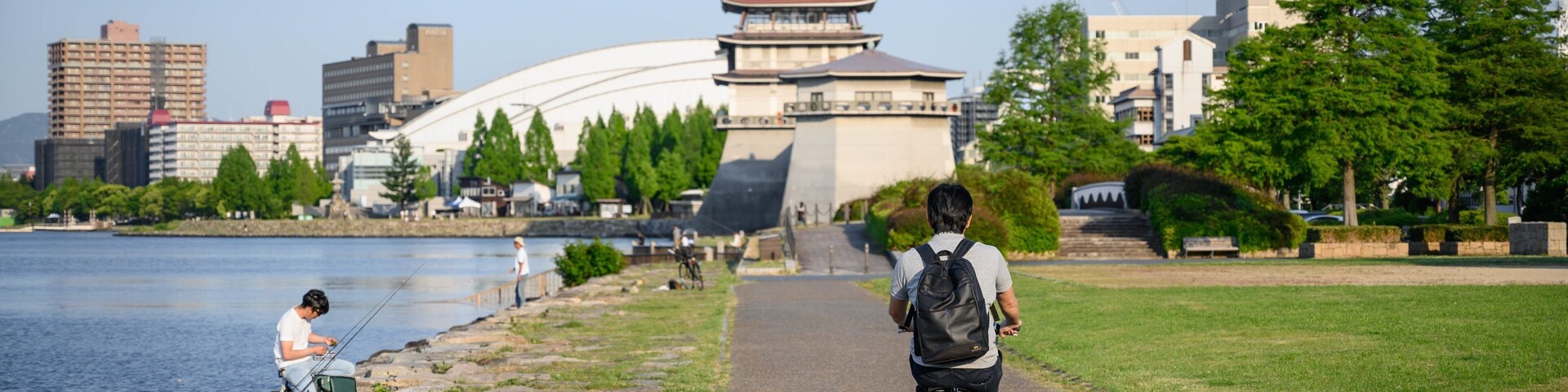 Una persona montando en bicicleta sobre un camino pavimentado junto a un río, con una pagoda tradicional al fondo.