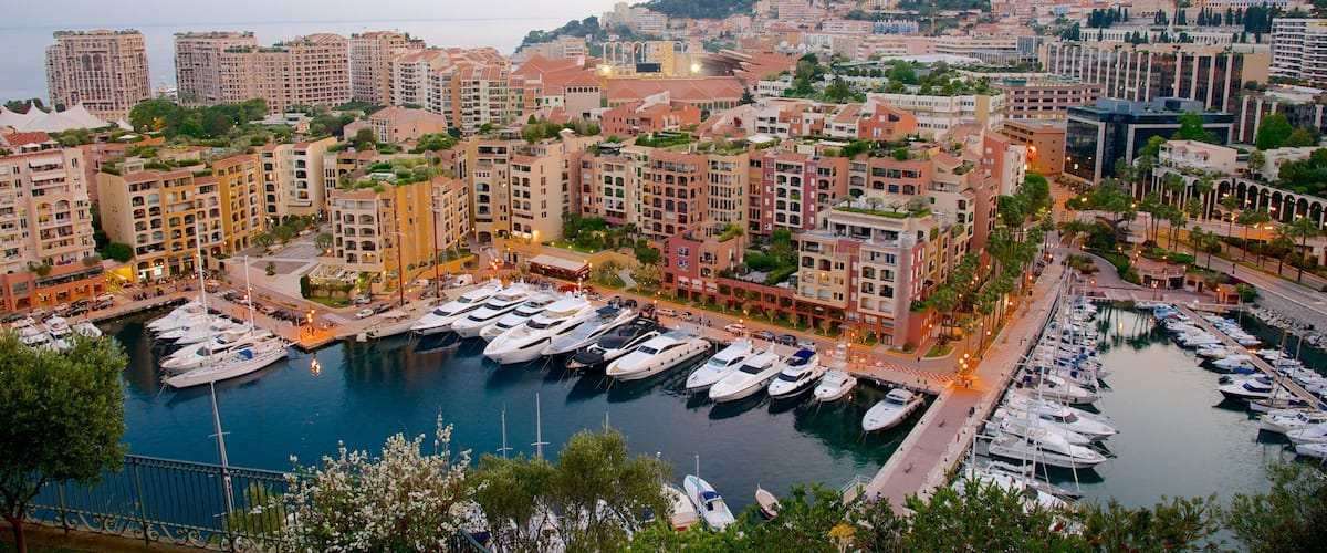 A marina with boats docked, surrounded by residential buildings and a mountainous backdrop.