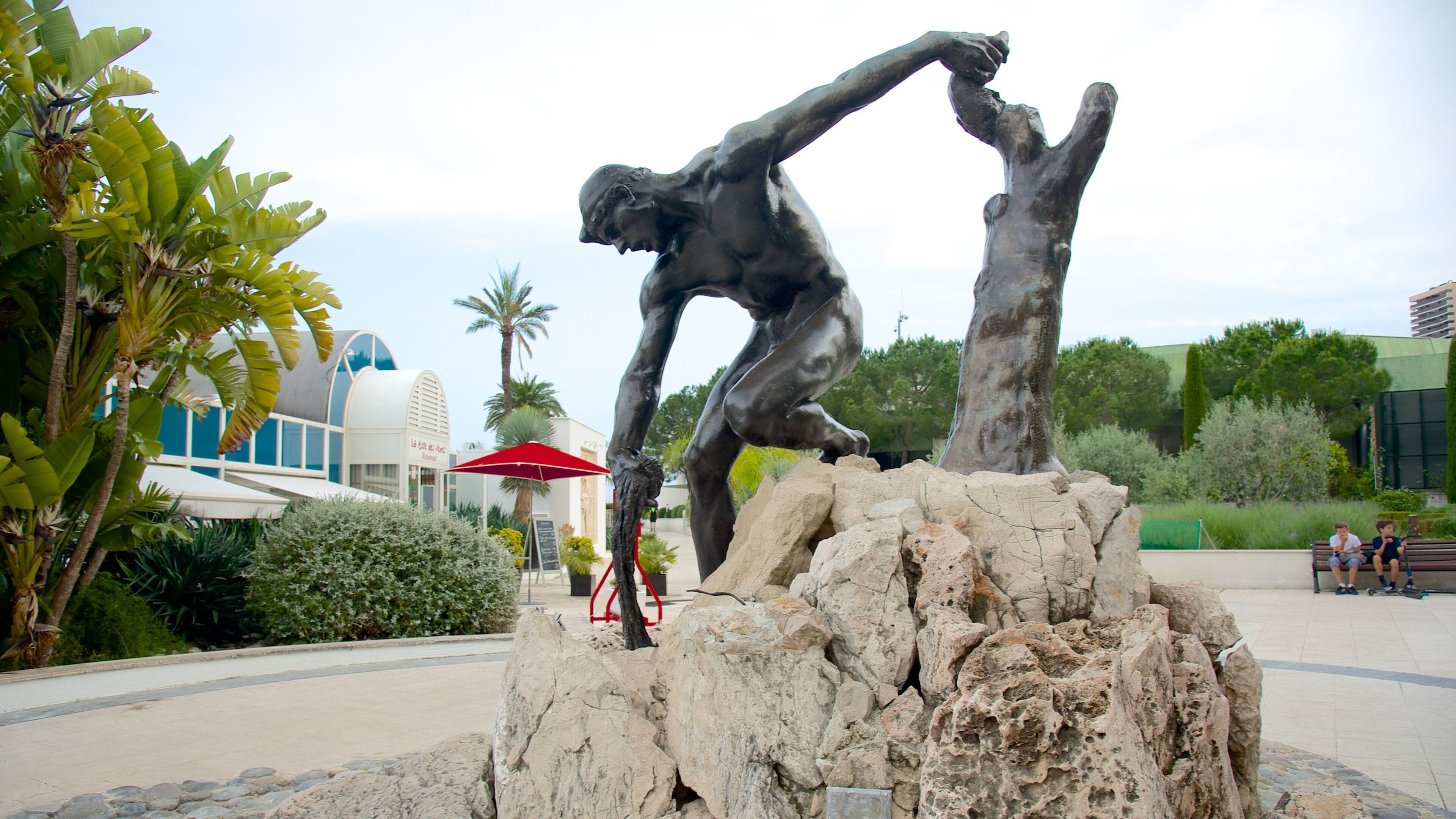 Una escultura de bronce de un hombre en una pose dinámica, con un fondo de plantas tropicales y un edificio moderno.