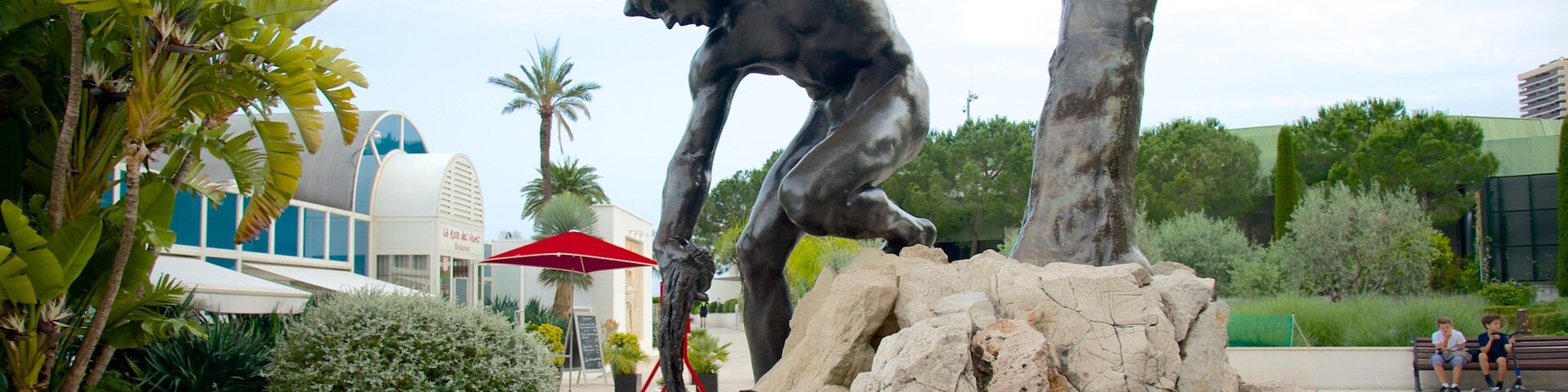Una escultura de bronce de un hombre en una pose dinámica, con un fondo de plantas tropicales y un edificio moderno.