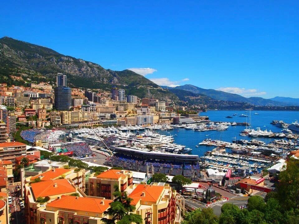 A coastal city with a marina filled with boats, surrounded by mountains and a mix of modern and historic buildings.