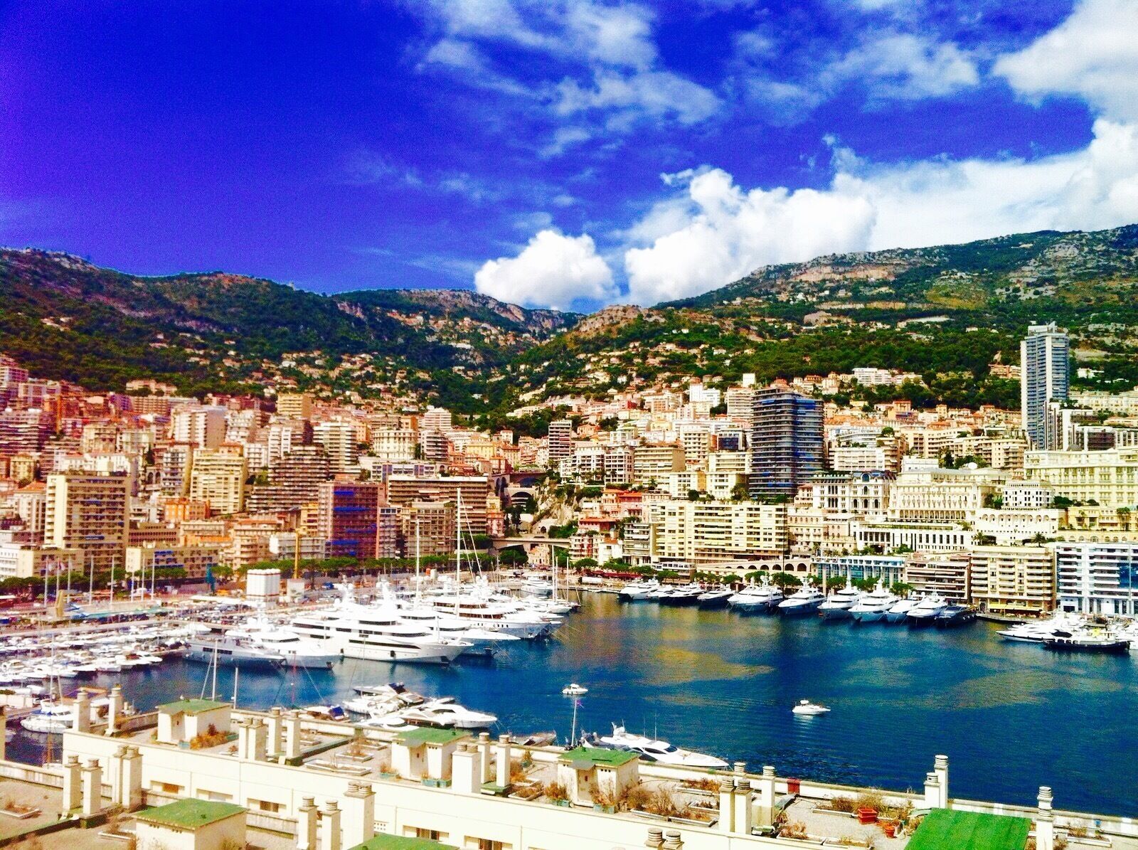 A coastal cityscape with a marina filled with yachts, surrounded by mountains and dense urban buildings.