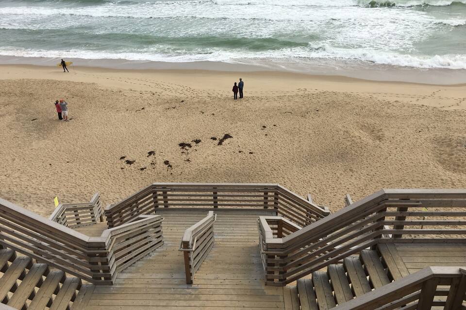 Un paseo marítimo de madera que conduce a una playa arenosa, con olas al fondo.