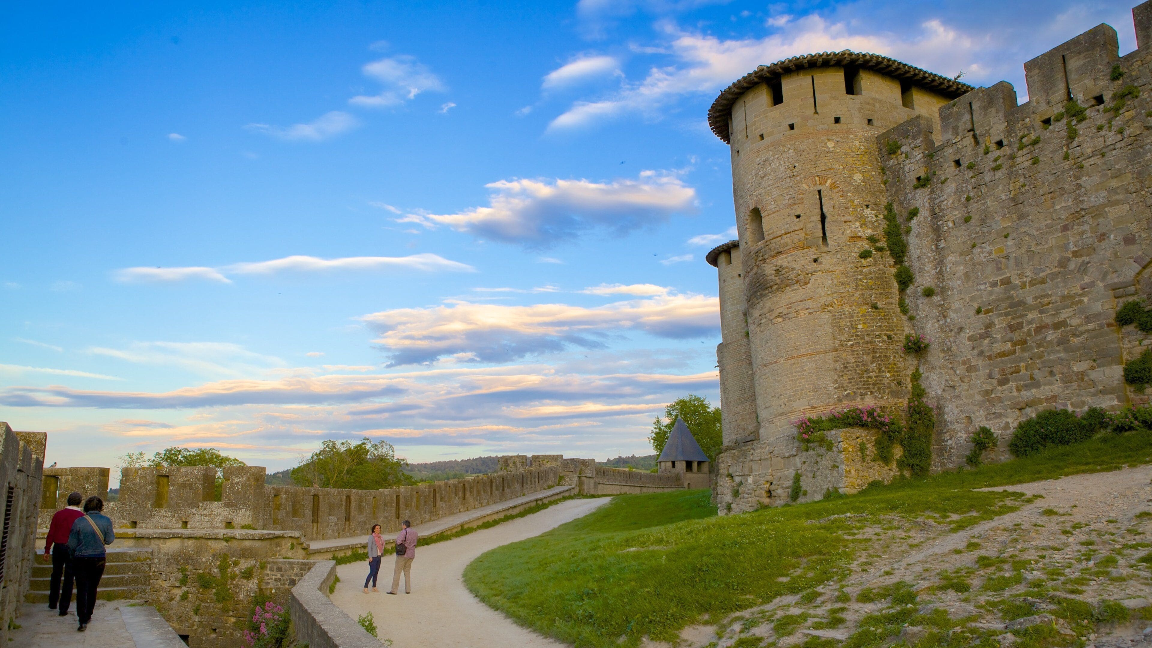 Un château historique en pierre, doté d’une haute tour ronde, d’un chemin pavé et d’une zone herbeuse.