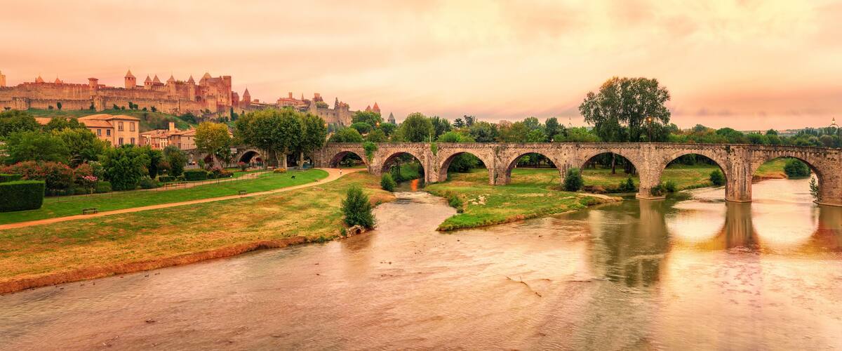 A stone bridge over a river, a historic castle in the background, and a park with a path.