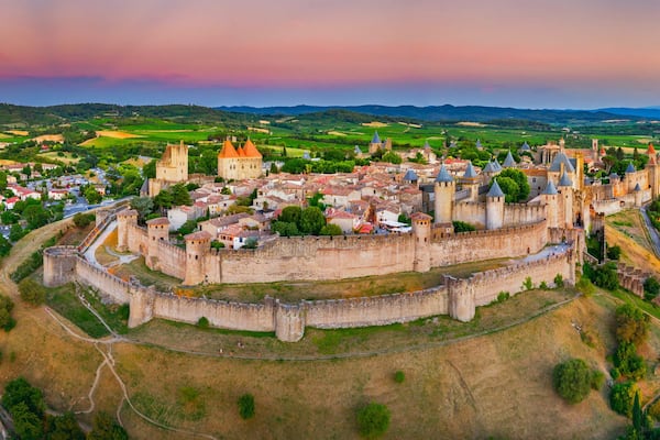 Un château médiéval entouré d’une ville fortifiée, avec des collines verdoyantes en arrière-plan.