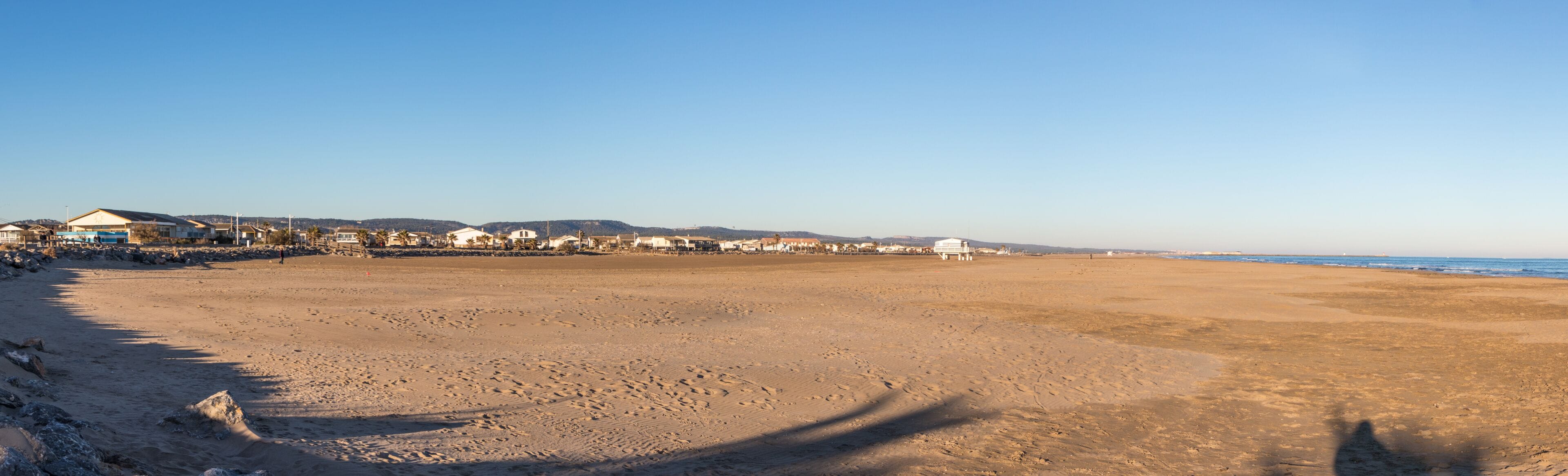 Una extensa playa arenosa con huellas, cielo azul despejado y edificios a lo largo de la costa.