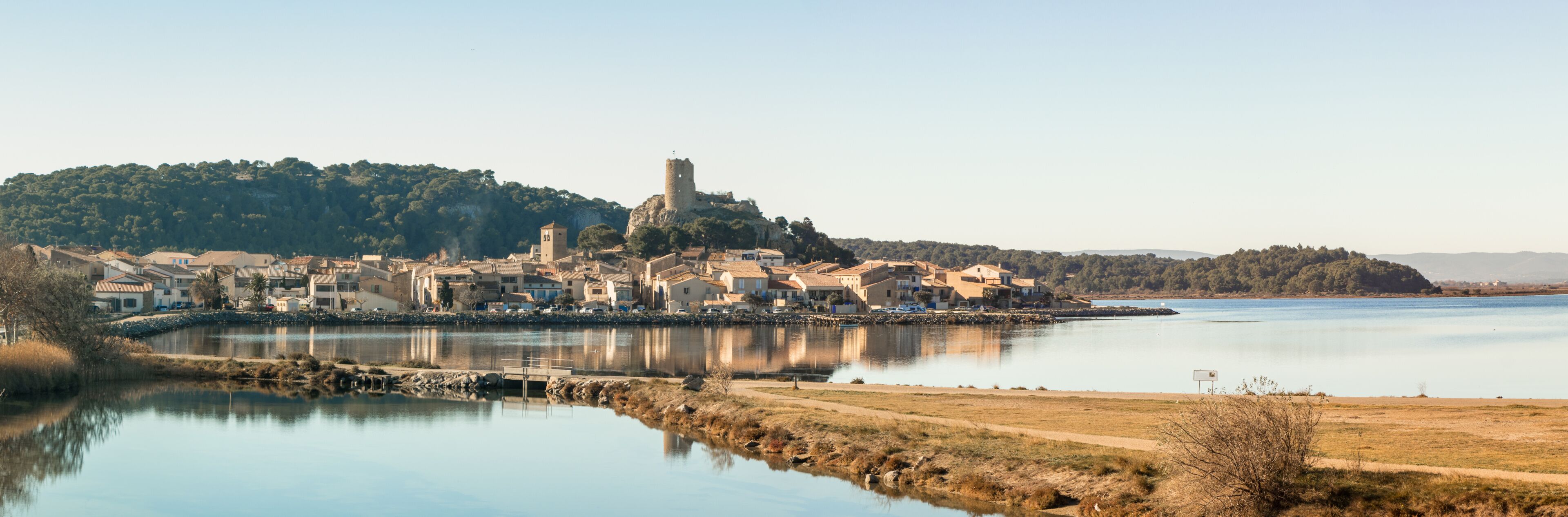 Uma vila costeira com um castelo imponente numa colina, águas calmas e um céu limpo.