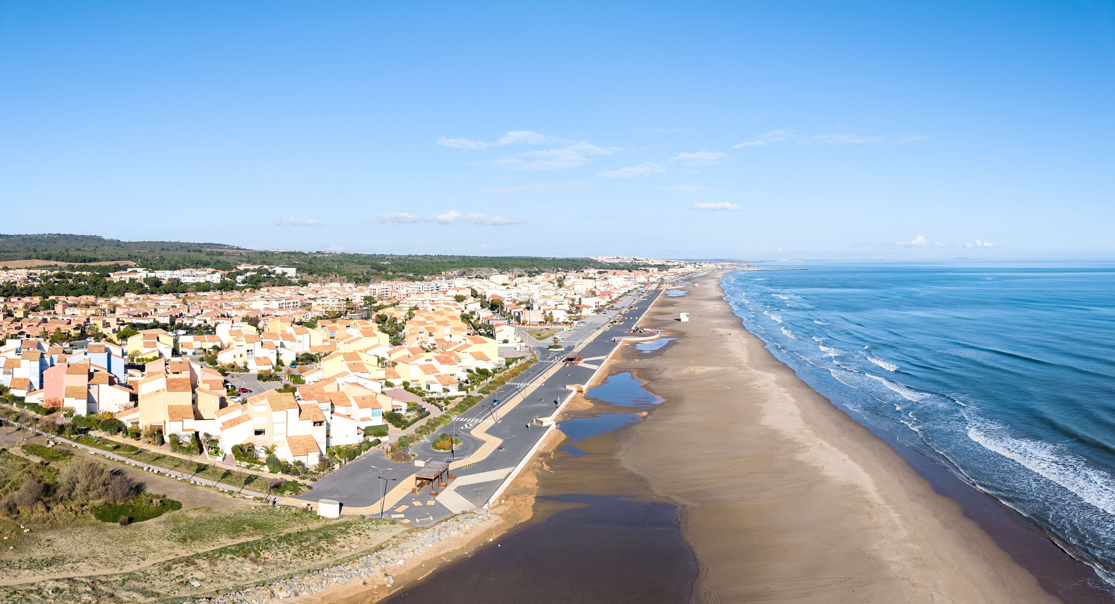 Un pueblo costero con una extensa playa, una carretera paralela a la orilla y edificios residenciales.