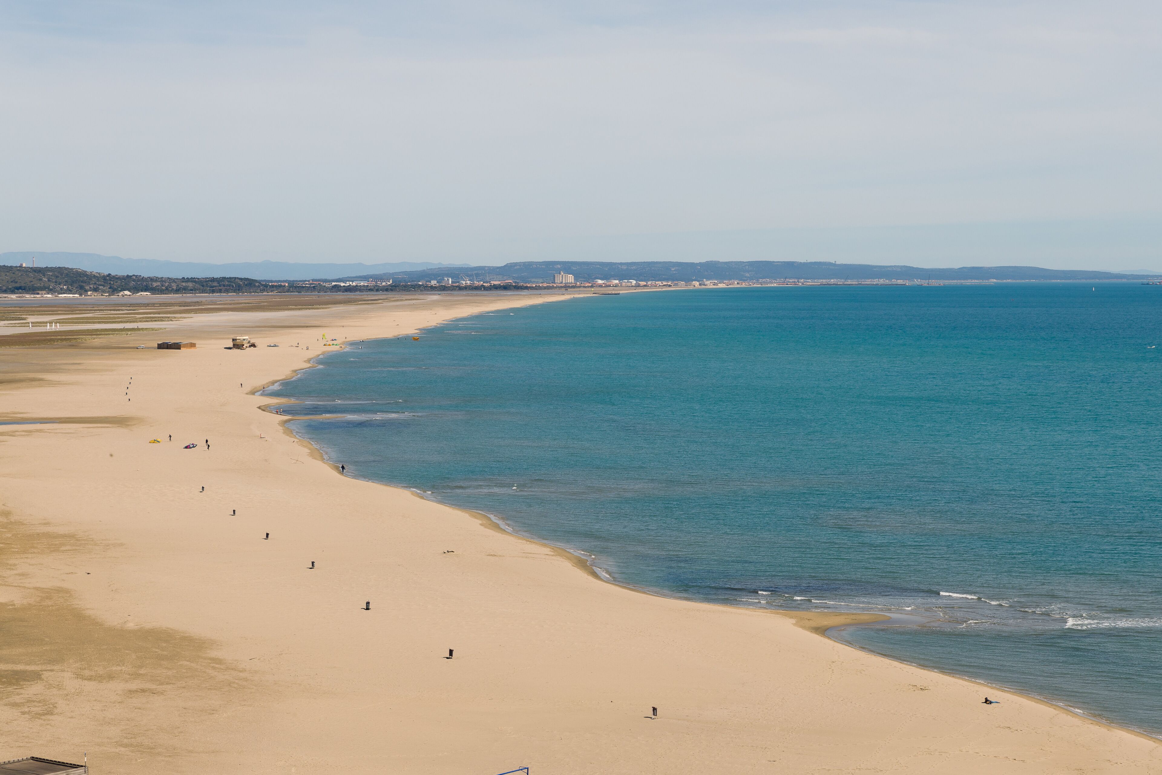 Una playa amplia y arenosa con un mar tranquilo, una silueta urbana distante y un cielo despejado.