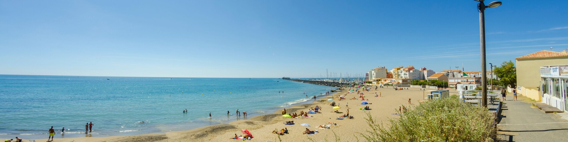 Una playa soleada con gente tomando el sol y nadando, un muelle que se adentra en el mar y edificios a lo largo de la costa.