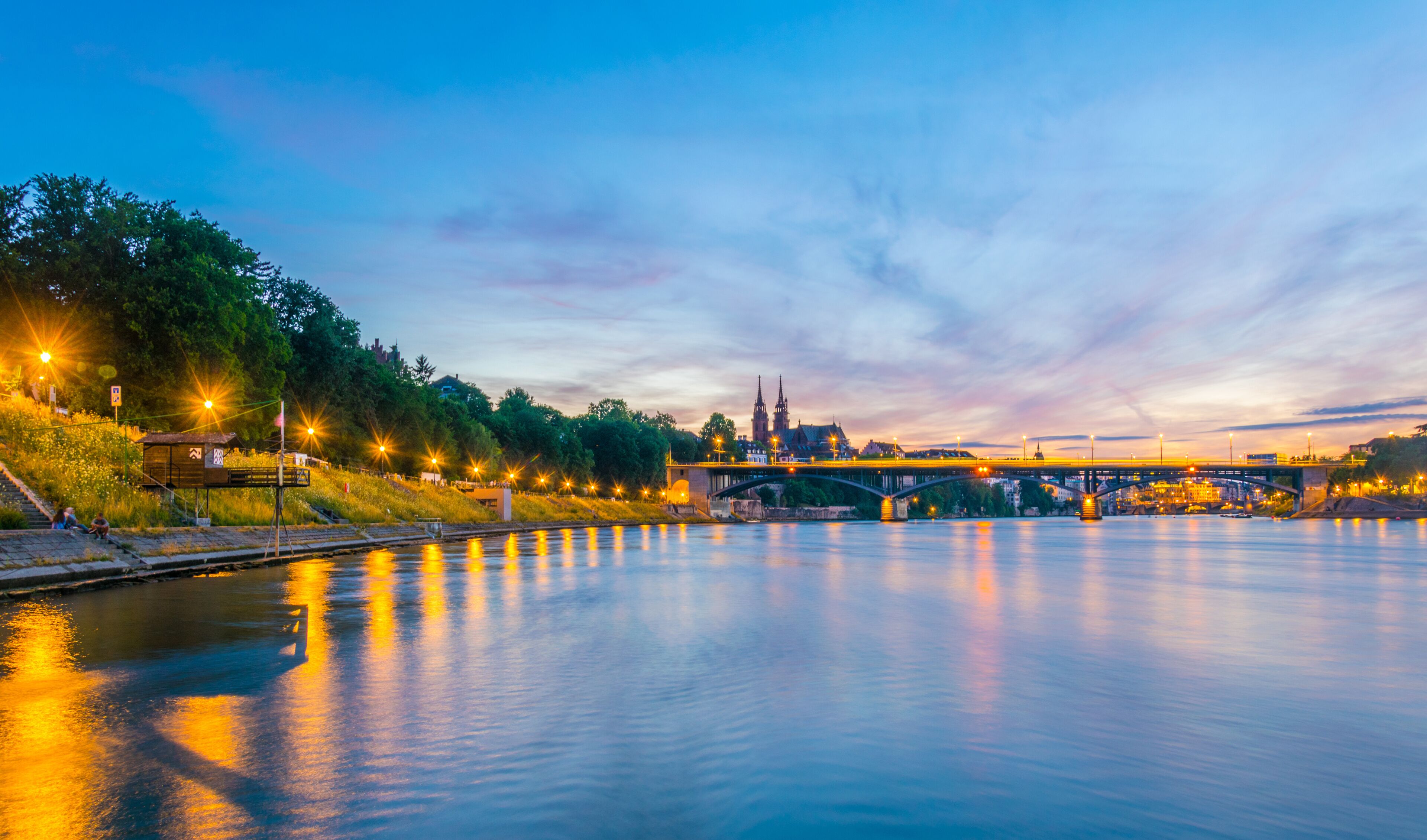A calm river with a bridge, a church, and a cityscape at dusk.