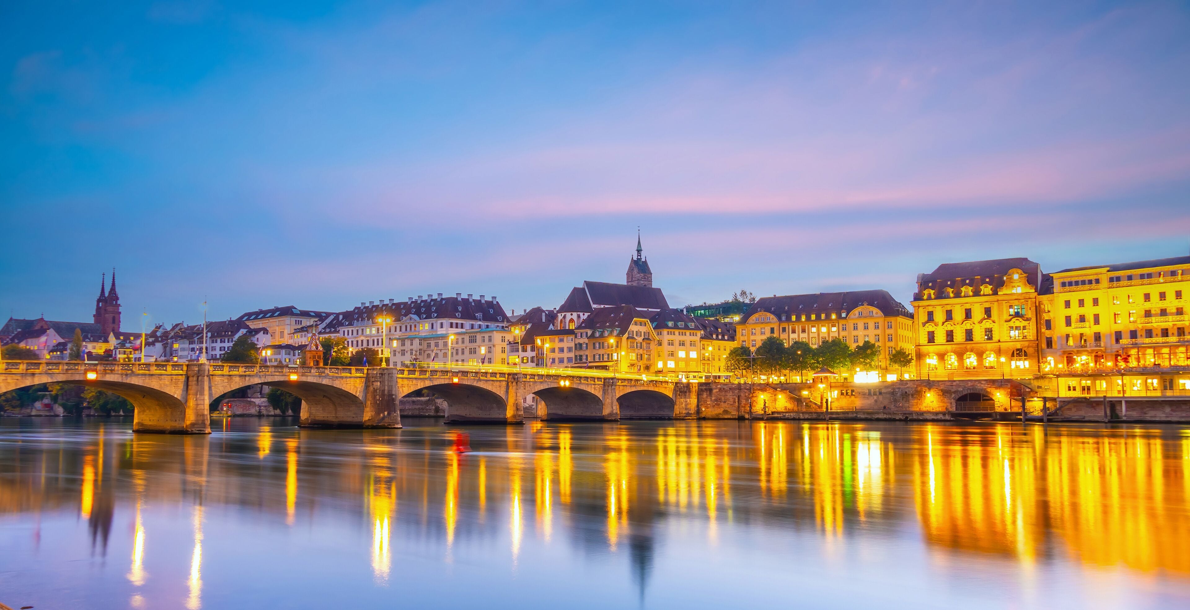 A river with a stone bridge, illuminated buildings, and a church spire.