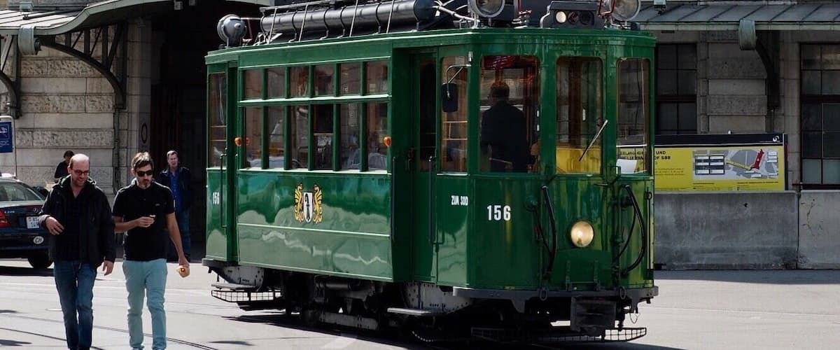 A green tram on tracks in front of a historic building with a clock and a sign reading "SBB CFF FFS".