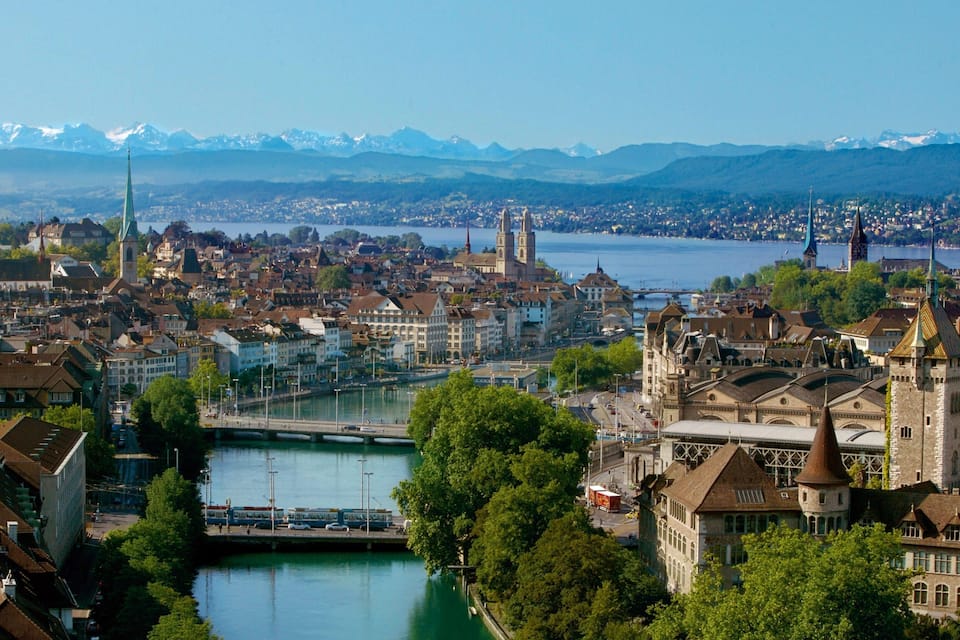 Ein weiter Blick auf eine Stadt mit einem Fluss, historischen Gebäuden und Bergen im Hintergrund.