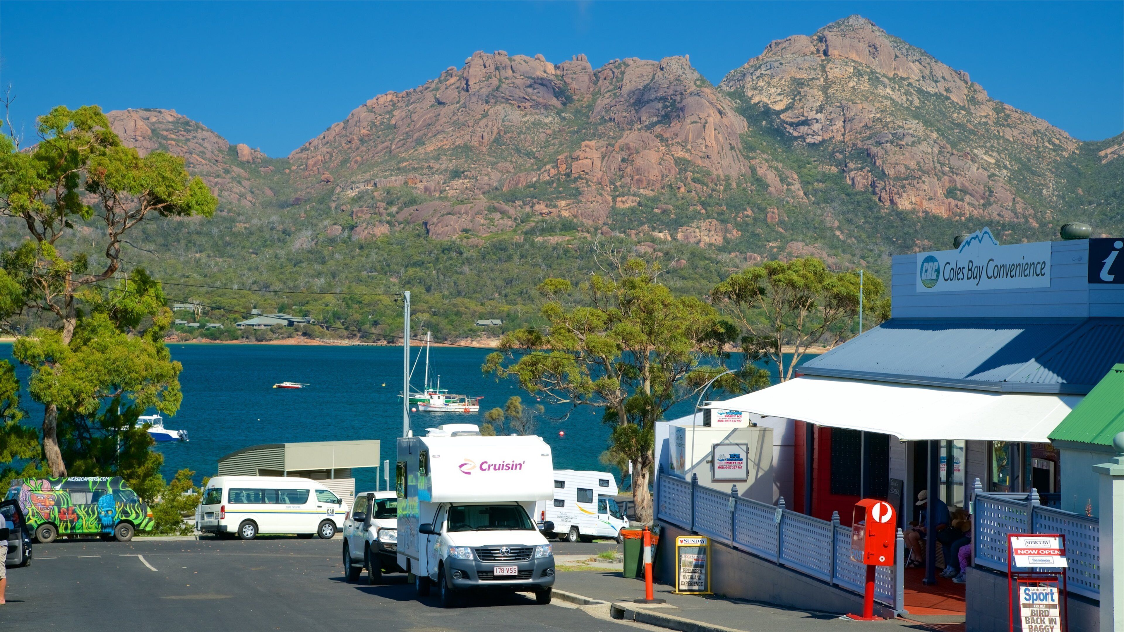 A coastal town with a marina, parked vehicles, and a mountain backdrop.