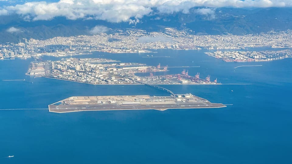 An aerial view of a coastal city with a large harbor, an airport, and mountainous terrain in the background.