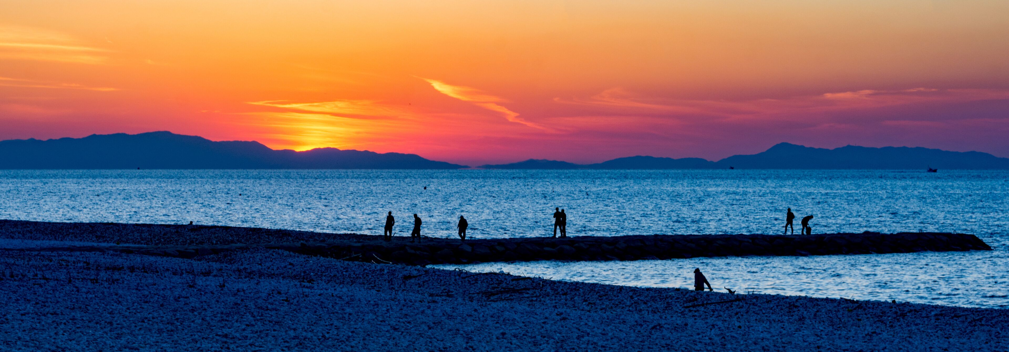 A beach at sunset with silhouettes of people and a pier extending into the water.