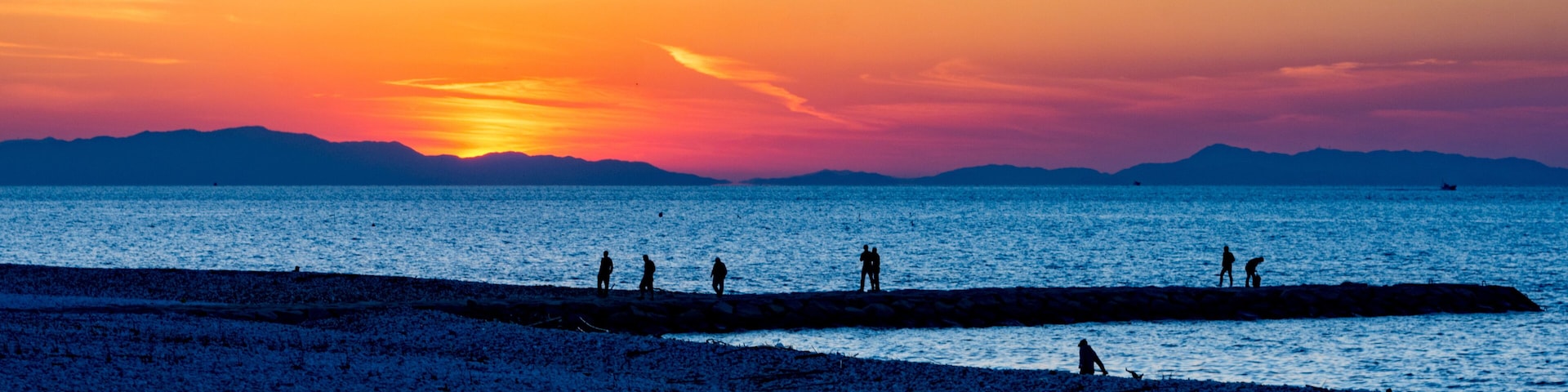 A beach at sunset with silhouettes of people and a pier extending into the water.