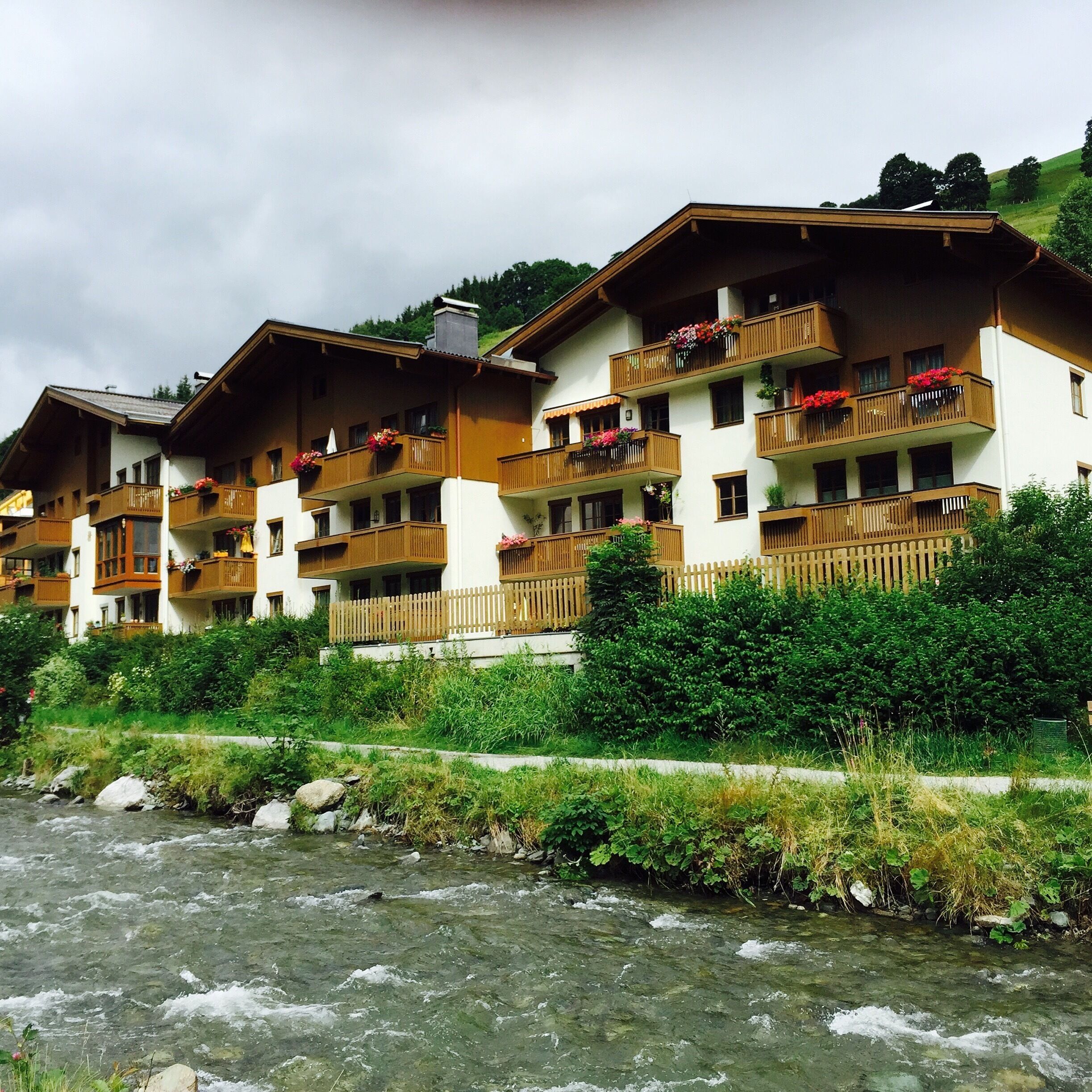 A row of chalet-style buildings with wooden balconies and flower boxes, situated beside a flowing river.