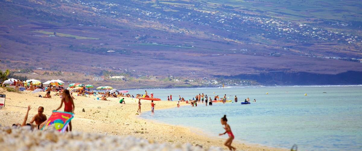 A beach with people sunbathing and swimming, surrounded by hills and houses.