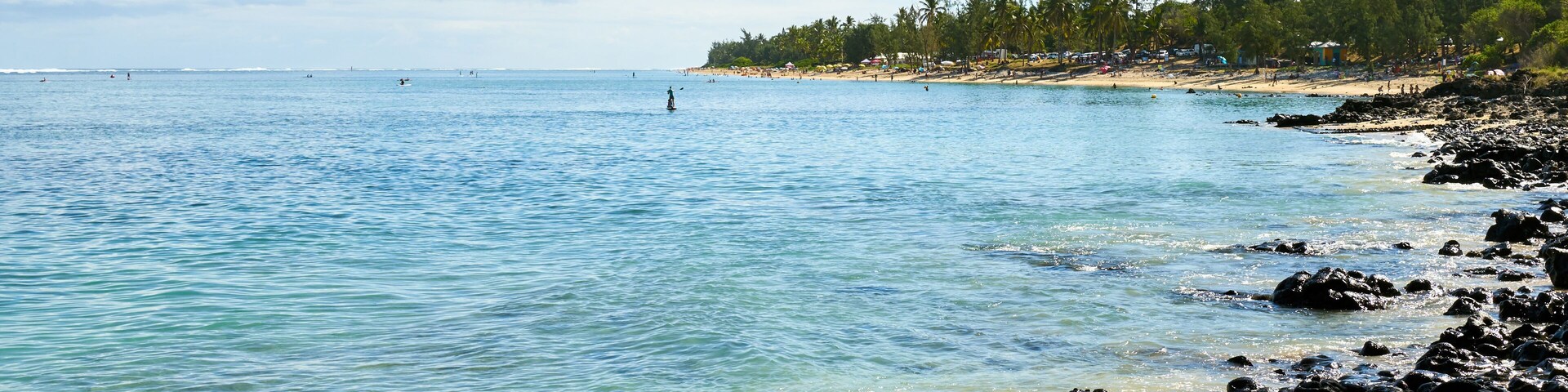 Una costa rocosa con agua azul cristalina, una playa arenosa y una fila de árboles al fondo.