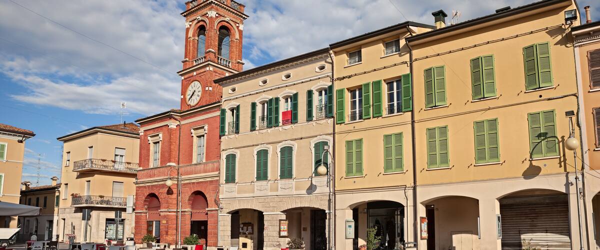 A historic square with a prominent clock tower, traditional buildings with green shutters, and outdoor seating areas.