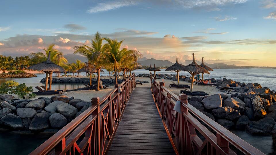 A wooden pier leading to a beach with palm trees and thatched umbrellas.