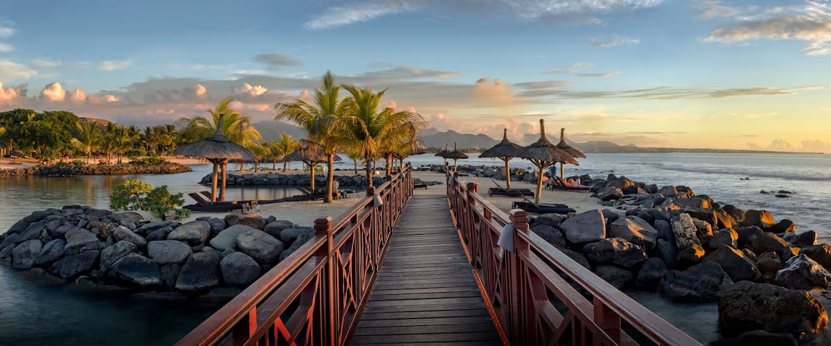 A wooden pier leading to a beach with palm trees and thatched umbrellas.