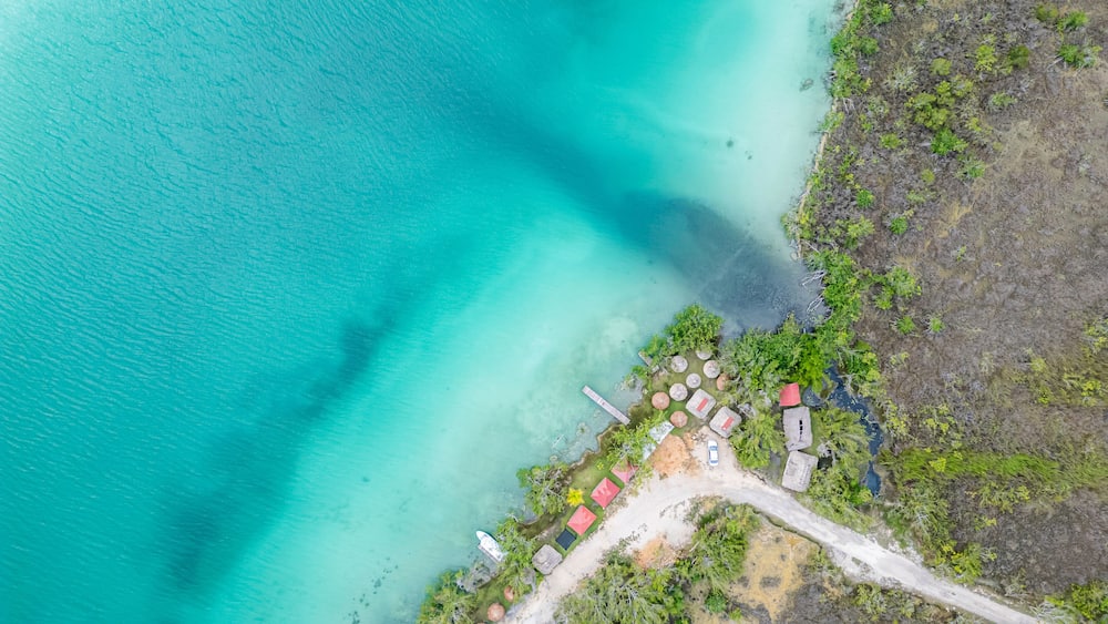 An aerial view of a coastal area with turquoise waters, a sandy beach, and several small structures with red roofs.