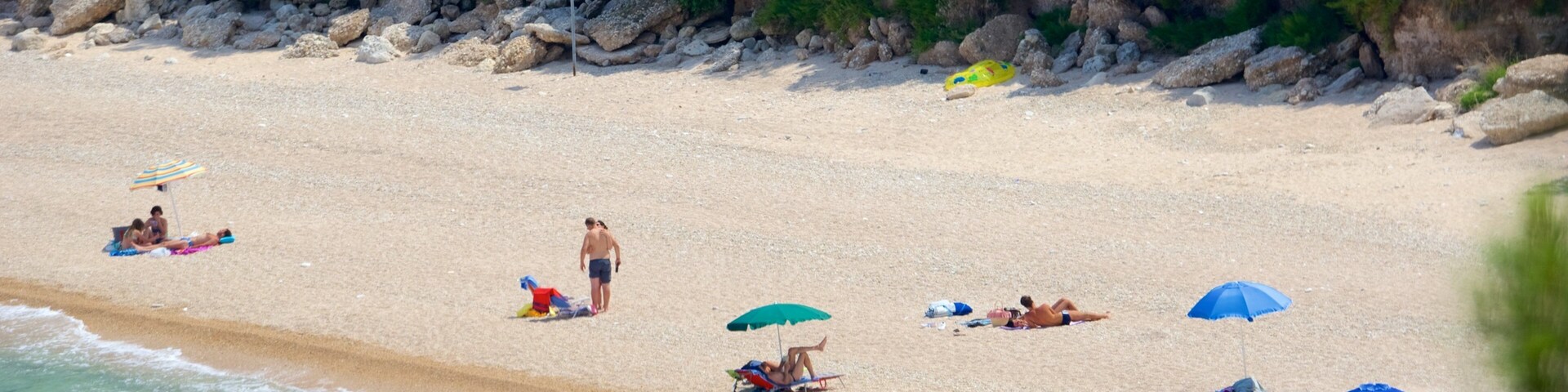 Una playa arenosa con gente tomando el sol, un bote y un acantilado rocoso al fondo.