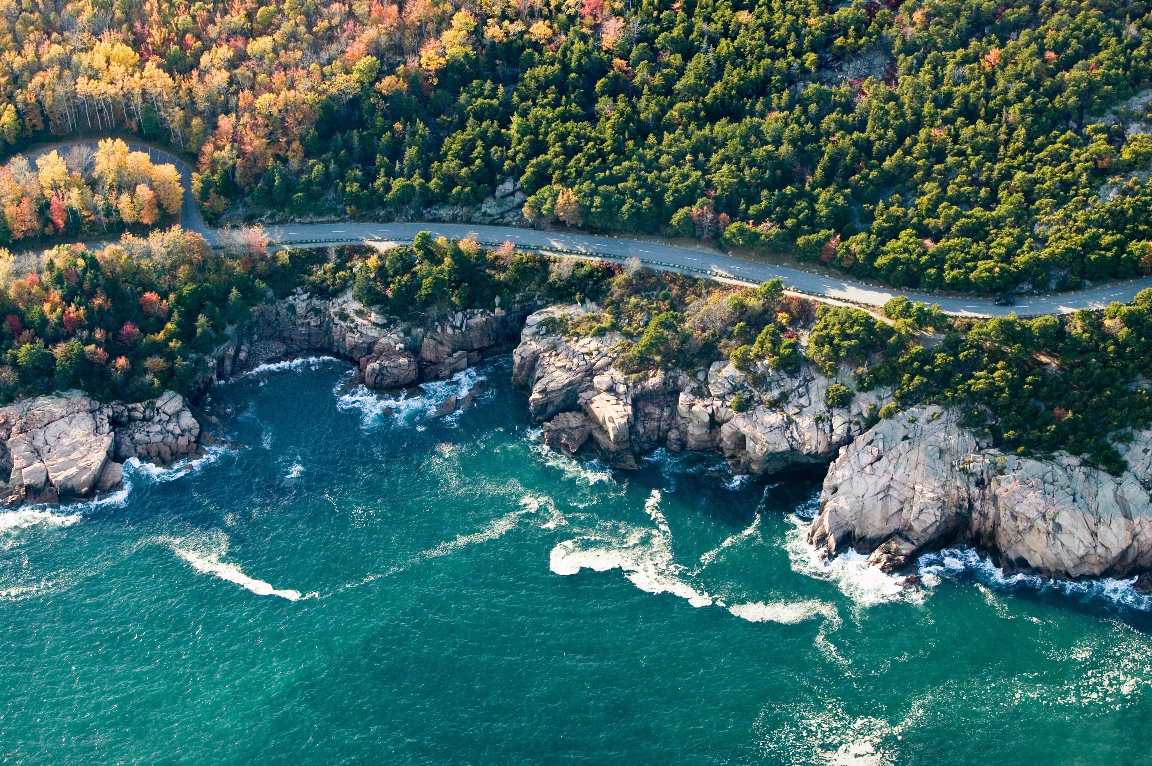 An aerial view of a coastal area with a winding road, rocky cliffs, and a forested landscape.