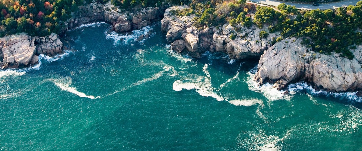 Vue aérienne d’une zone côtière avec une route sinueuse, des falaises rocheuses et un paysage boisé.