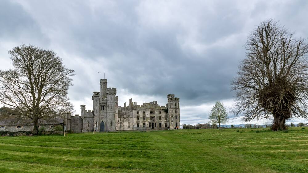 A historic castle with multiple towers and turrets, surrounded by a grassy field and leafless trees under a cloudy sky.
