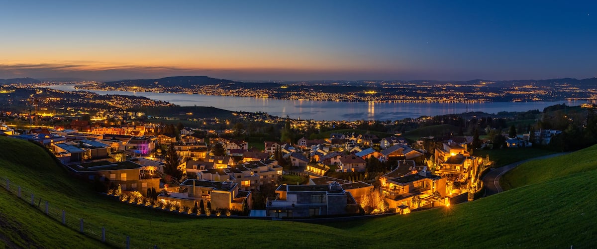 A coastal town at dusk with a river, hills, and illuminated buildings.
