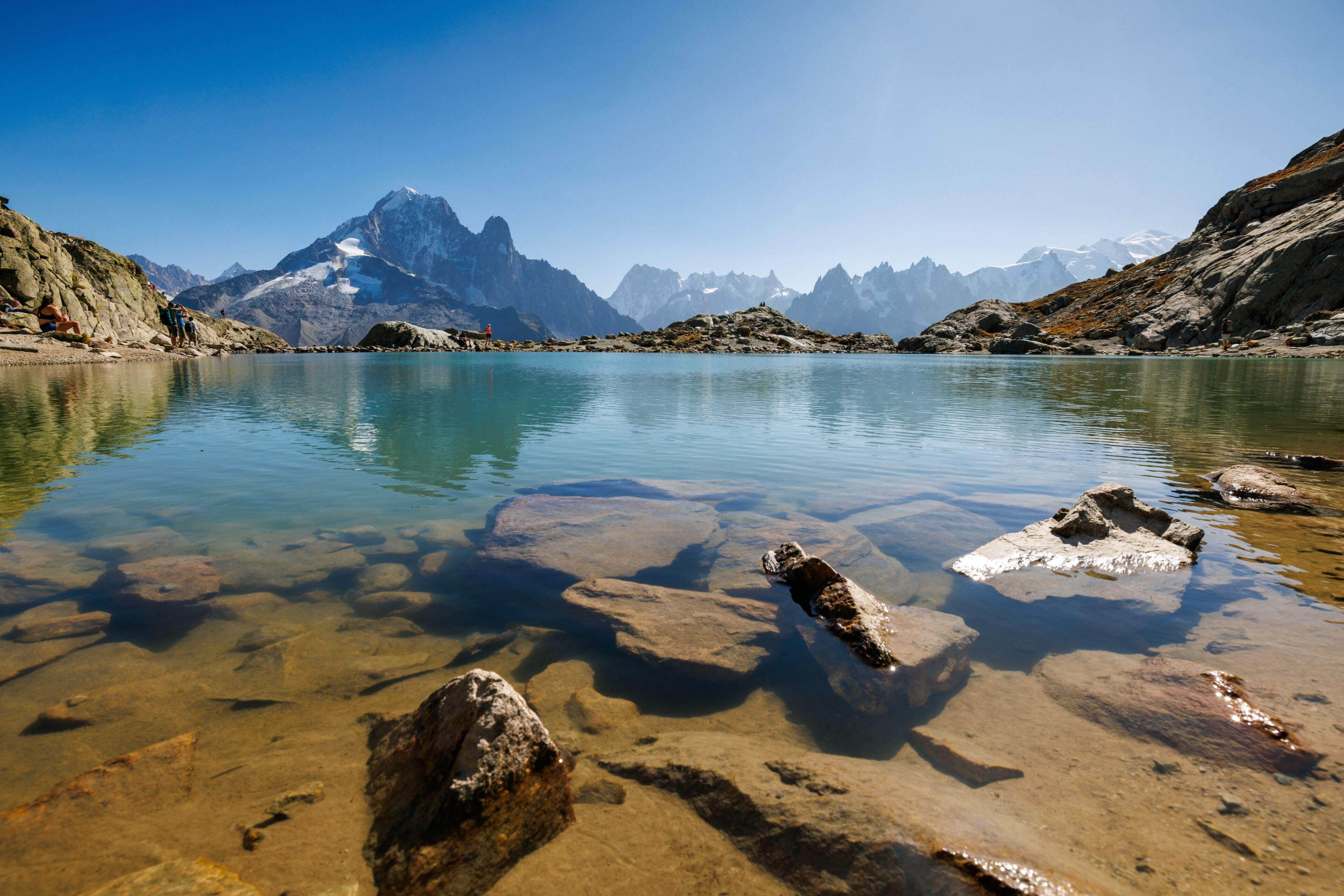 Um lago límpido com pedras visíveis e uma cordilheira ao fundo.