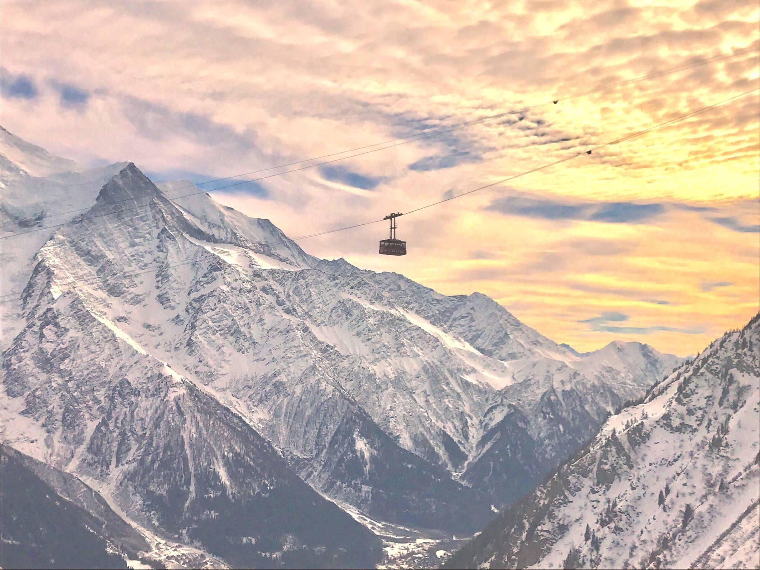 Une télécabine se déplace au-dessus d’une chaîne de montagnes enneigées, avec un ciel embrasé par le coucher du soleil.