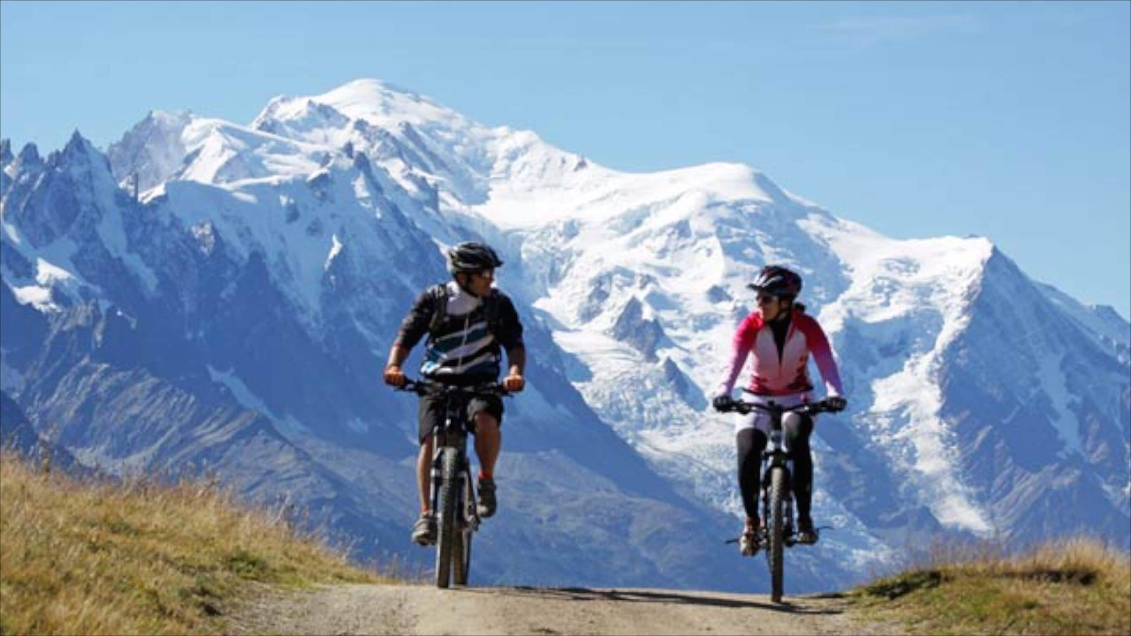 Two cyclists on a mountain road with snow-capped peaks in the background.